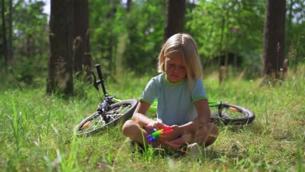 Rubia Boy se sienta en un parque en tiempo soleado y juega pop-it — Vídeos de Stock