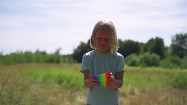 Blondýny chlapec v parku hraje pop-it Rainbow barvy. — Stock video