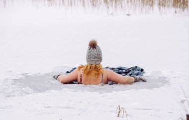 Yün şapka takan, eldiven takan, mayo giyen, göl kenarındaki ev yapımı buz deliğinde yüzen bir kadın. Sağlıklı kış buz banyosu konsepti.