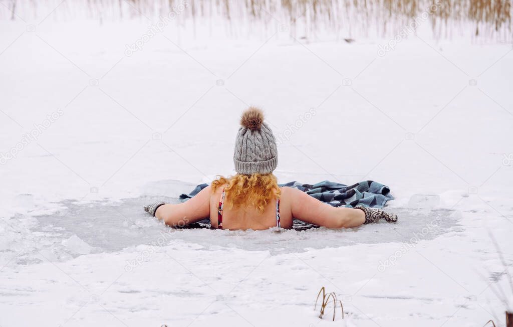 Woman wearing wool hat, mittens gloves and swimsuit, swimming dipping inside homemade ice hole in lake. Healthy winter ice bath concept.