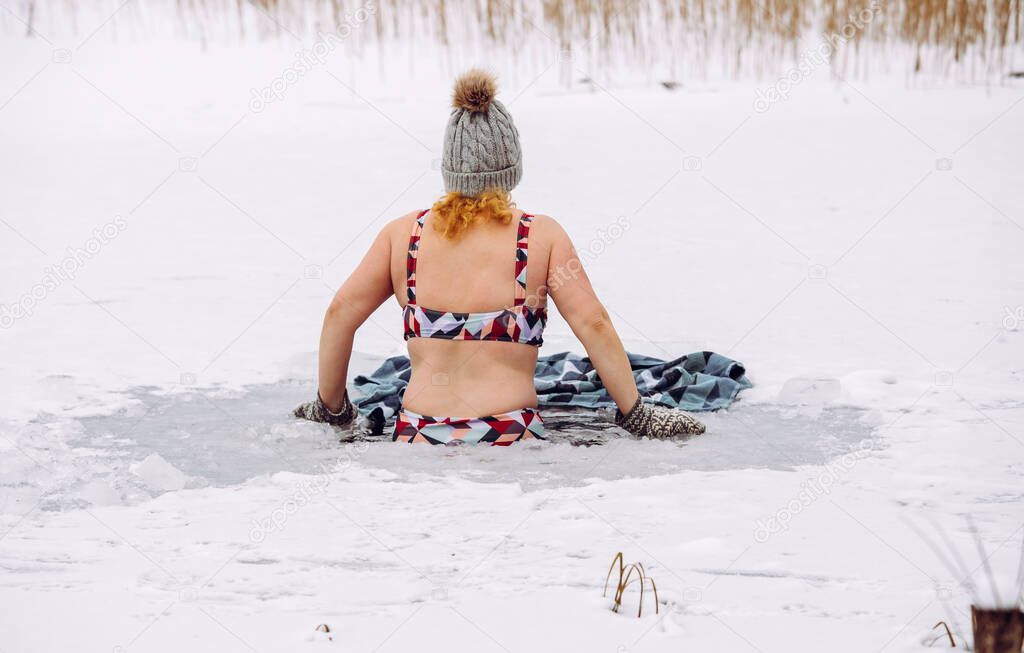 Woman wearing wool hat, mittens gloves and swimsuit, swimming dipping inside homemade ice hole in lake. Healthy winter ice bath concept.
