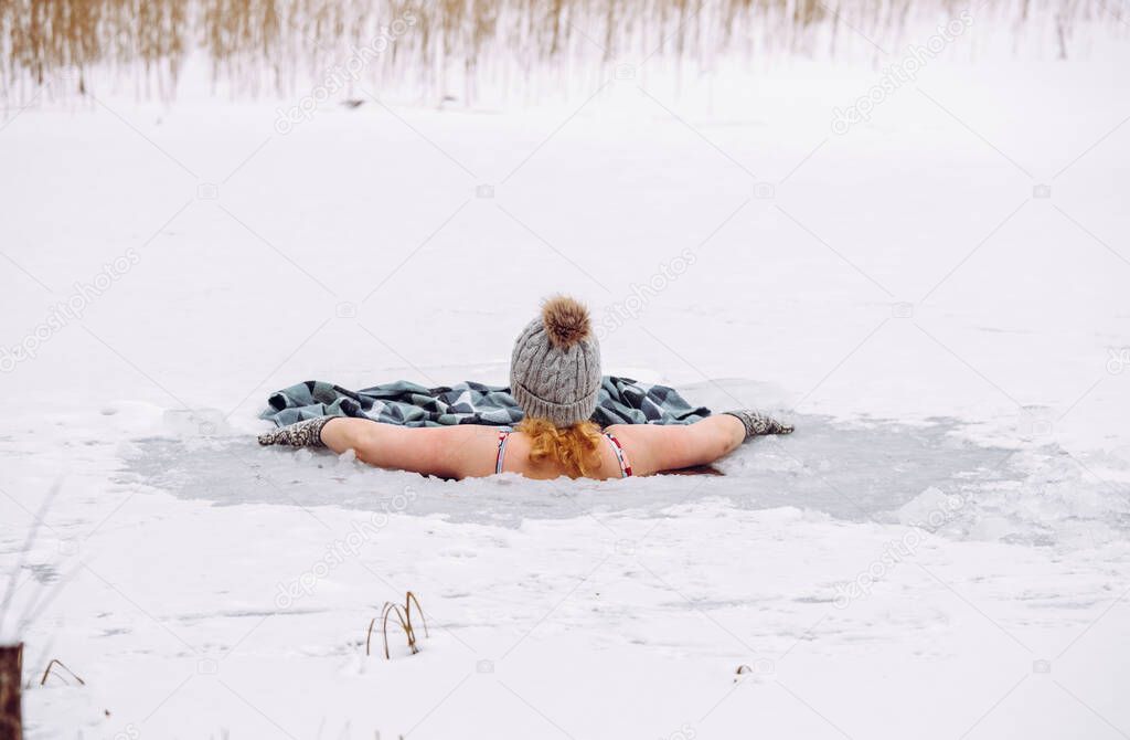 Woman wearing wool hat, mittens gloves and swimsuit, swimming dipping inside homemade ice hole in lake. Healthy winter ice bath concept.