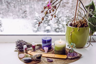 Small good feng shui altar in home on window sill on leaf shape table mat, snowy Nordic nature on background. Incense candle smoking, gemstones and orchid flower pot. clipart