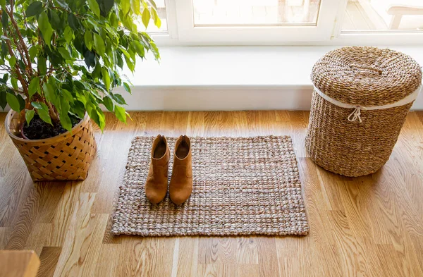 Hardwood floor with jute doormat, shoes and flower pot and seagrass laundry basket by window. Natural material objects in home concept. Home interior.