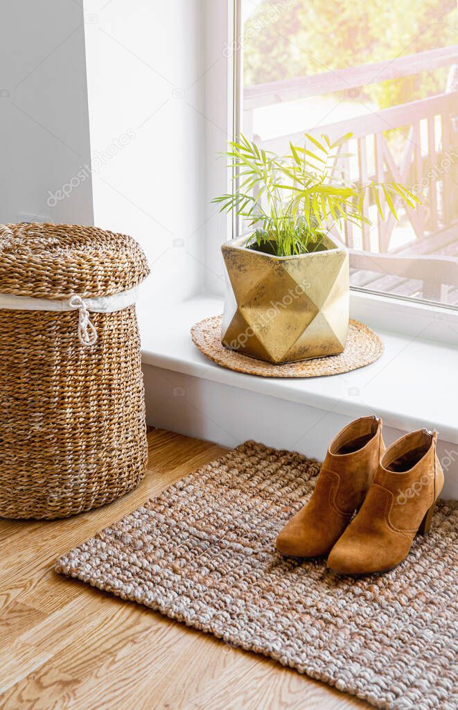 Hardwood floor with jute doormat, shoes and flower pot and seagrass laundry basket by window. Natural material objects in home concept. Home interior. 