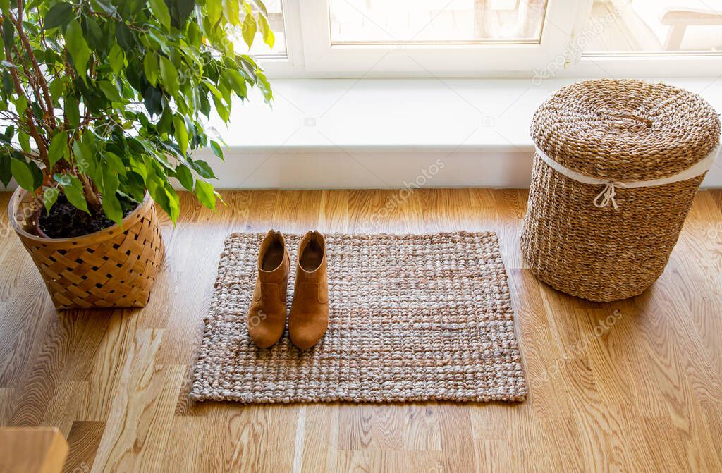 Hardwood floor with jute doormat, shoes and flower pot and seagrass laundry basket by window. Natural material objects in home concept. Home interior. 