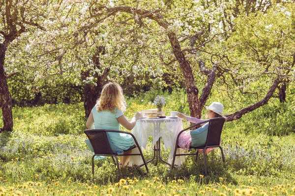 Mother daughter spending time together in garden under the blooming spring apple trees, drinking tea and spending time together. Idyllic sunny day setting.