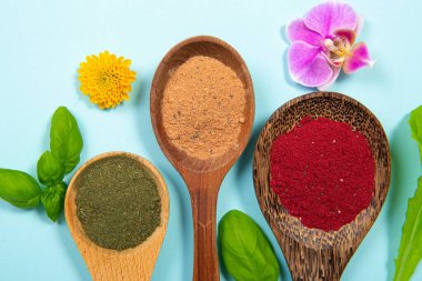 Çiçek çiçekleriyle süslenmiş kaşığın üzerinde çeşitli süper yiyecek tozları ve mavi stüdyo arka planında yapraklar vardı. Canlı renkli resim kırmızı, yeşil, sarı, mavi.