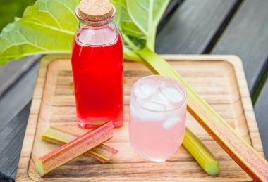 Homemade rhubarb syrup ( Rheum rhabarbarum ). Nice pink liquid syrup in bottle and glass with juice and ice cubes in drinking glass on tray, decorated with rhubarb stalks. Refreshing spring drink. clipart