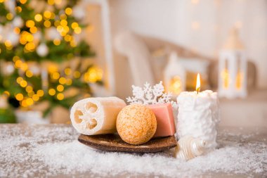 Kış spa şımartma konsepti. Çeşitli kişisel bakım ürünleri banyo bombası, sabun, ahşap tepside luffa süngeri mum yakma ve arka planda fenerler olan Noel ağacı..