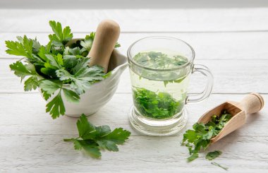 Maydanoz Petroselinum gevreğinden yapılmış ev yapımı şifalı bitki çayı. Yeşil yapraklar çay fincanında beyaz ahşap zemin üzerinde taze havan ve tokmak yapraklarıyla.