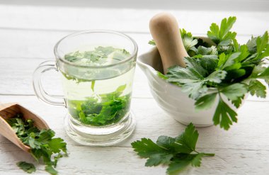 Maydanoz Petroselinum gevreğinden yapılmış ev yapımı şifalı bitki çayı. Yeşil yapraklar çay fincanında beyaz ahşap zemin üzerinde taze havan ve tokmak yapraklarıyla.