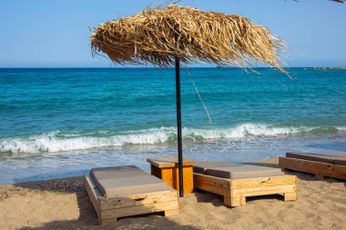 Modern ahşap yastıklı güneşlenme sandalyeleri, deniz kenarında, kamış güneş şemsiyelerinin altında, güzel mavi bir deniz ve yazları mavi gökyüzü. Boşluğu kopyala.