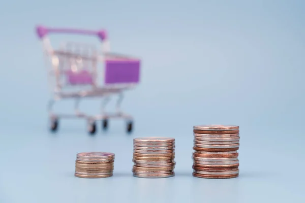 Coin stacks with empty shopping cart on blue background. Increasing shopping expenses concept