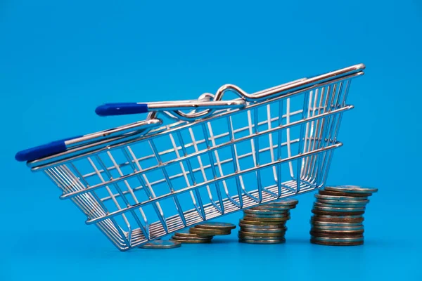 Empty shopping basket on coin stacks on blue background. Growing shopping expenses concept