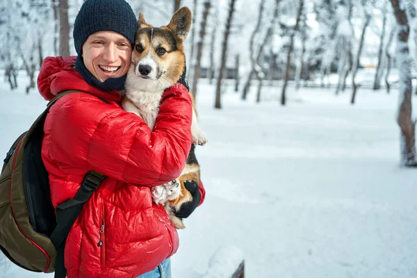 Winter portrait man wearing warm clothes embracing his pet Corgi dog in winter at snow park — Stock Photo, Image