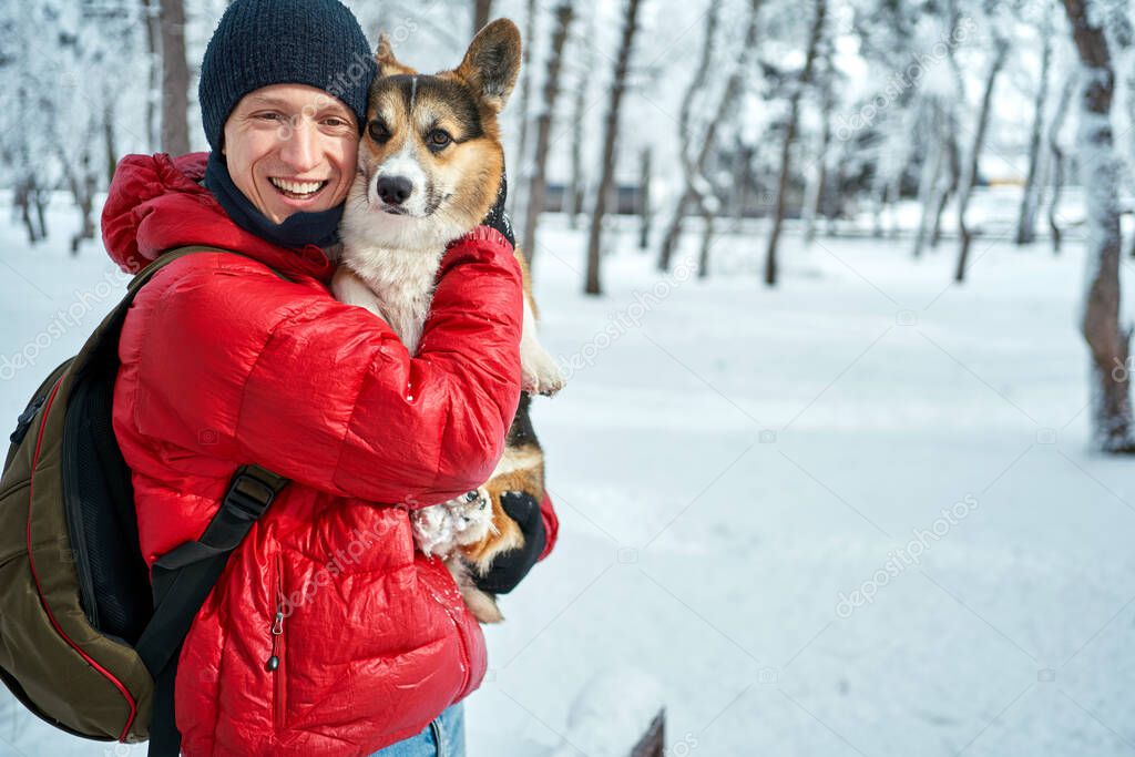 winter portrait man wearing warm clothes embracing his pet Corgi dog in winter at snow park