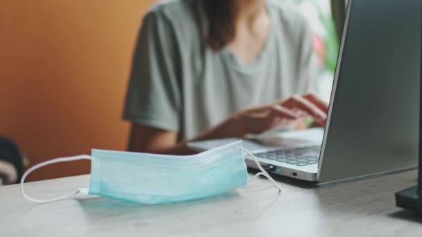 Utilizado máscara médica protectora está en la mesa en el lugar de trabajo y la mujer usando el ordenador portátil — Vídeo de stock