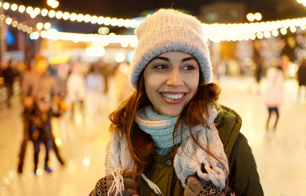 Zbliżenie portret cute girl w czapce zimowej na lodowisku tle z świąteczne lampki na wieczór. — Zdjęcie stockowe