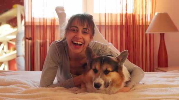 Close-up portrait of pleased woman lying on bed at home with her loved dog Welsh Corgi. — Stock Video