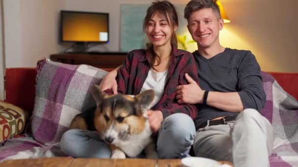 Happy couple with cute pet Welsh Corgi dog sitting on couch at home at evening — Stock Video