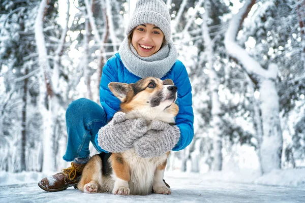 Zima uśmiechnięta kobieta w ciepłym kapeluszu i szaliku przytula się do swojego zwierzaka Welsh Corgi doga w śniegu w zimowym parku — Zdjęcie stockowe