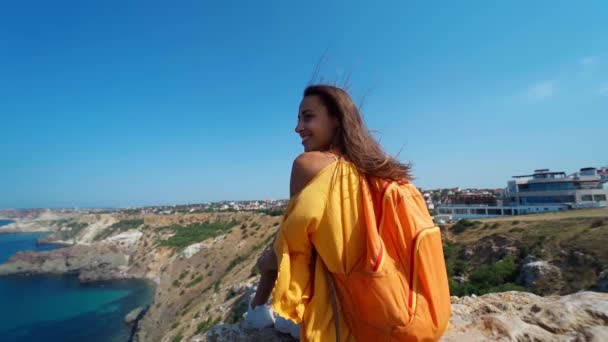 Vista trasera hermosa mujer viajero en vestido de naranja sentado en el borde del acantilado — Vídeo de stock