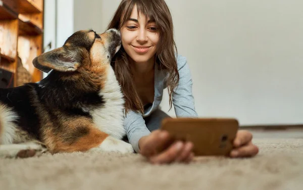 Femeie întinsă pe podea cu animale de companie și făcând fotografie selfie, câine galez Corgi sărutându-și proprietarul — Fotografie, imagine de stoc