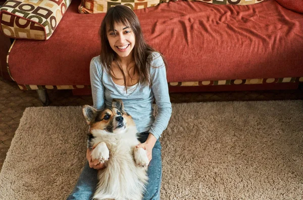 Mujer encantada sentada en la alfombra, jugando y acariciando perro galés Corgi. amistad con la mascota y el propietario —  Fotos de Stock