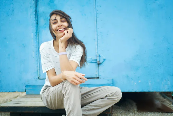 Nette glückliche unbeschwerte Frau sitzt auf der Veranda vor alter blauer Wand und posiert lächelnd in die Kamera — Stockfoto