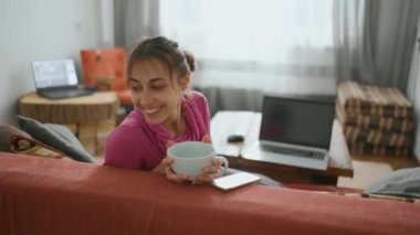 Neşeli kız elinde büyük bir fincan çayla evinde oturuyor.