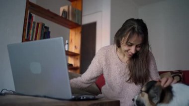 Kanepede dizüstü bilgisayarıyla çalışan ve köpeğini okşayan bir kadın.
