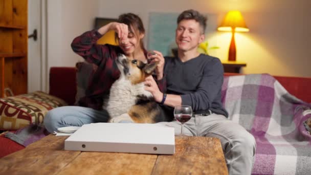 Familia en el sofá acariciando y alimentando perro, viendo la televisión y riendo — Vídeos de Stock