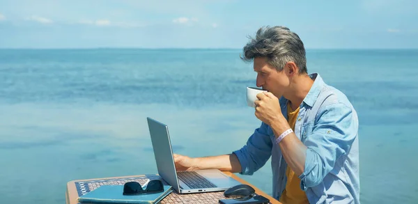 En tankefull frilanser som jobber på en bærbar datamaskin ved stranden, drikker kaffe. stockbilde