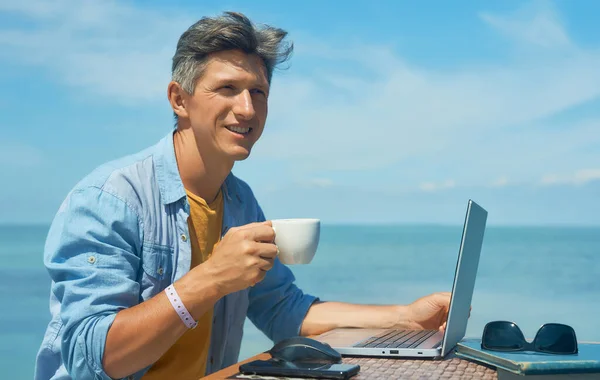 Portret optimist om zâmbitor freelancer care lucrează în aer liber pe plajă de mare albastră, bea cafea Imagini stoc fără drepturi de autor