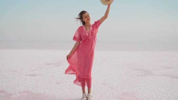 Kvinna i blåsa airness rosa klänning och hatt stående på salta stranden på rosa sjö — Stockvideo