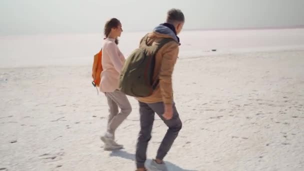Šťastný krásný pár miléniální hipsteři chůze ruku v ruce a radostně běží na růžové bílé slané pobřeží — Stock video