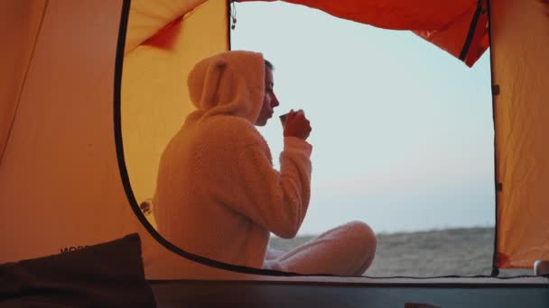 Achteraanzicht van mooie vrouw in grappige pyjama als konijn zitten bij de ingang van de tent en het drinken van hete koffie uit metalen mok — Stockvideo