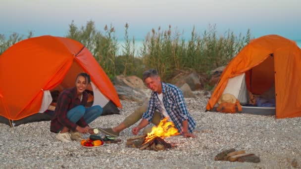 Giovane coppia amorevole di turisti campeggio, seduto vicino al fuoco contro tende arancioni sulla spiaggia — Video Stock
