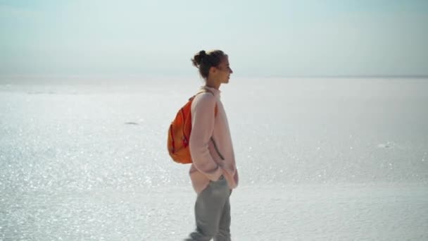 Radosna kobieta w przytulnym swetrze z pomarańczowym plecakiem na naturalnym krajobrazie słonych mieszkań, wygląda jak biała pustynia — Wideo stockowe