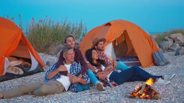 Två söta par omfamnar varandra och sitter vid brasan i havet camping på kvällen och tittar på havet — Stockvideo