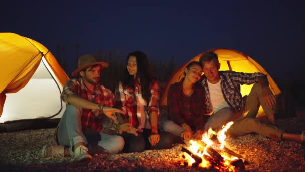 Amigos relaxando, aquecendo e se divertindo pela fogueira no acampamento com tendas na praia — Vídeo de Stock