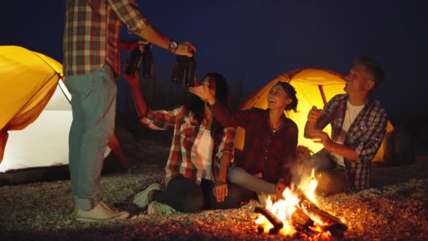 Hombre joven adulto en ropa casual trae cuatro botellas de cerveza a sus amigos en la hoguera en la playa acampando con tiendas de campaña — Vídeos de Stock