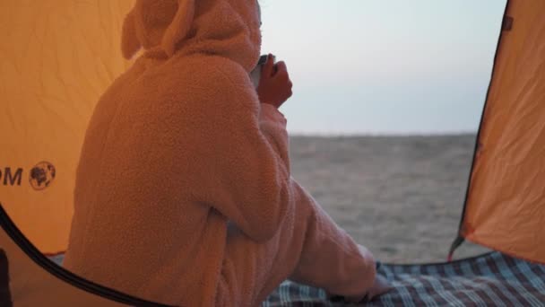 Bakifrån av söt kvinna i rolig pyjamas som kanin sitter vid tält inträde och dricka varmt kaffe från metall mugg — Stockvideo