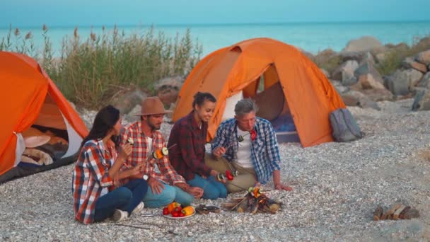 Sekelompok orang dewasa yang riang tiba di tepi laut pada sore hari sambil makan sayuran panas yang dipanggang di dekat api — Stok Video