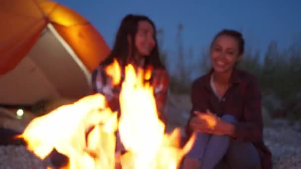 Ateşin önünde oturmuş şakalaşan ve kamp ateşine gülen iki kız arkadaş. — Stok video
