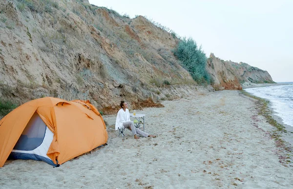 Sommerstrand-camping. kvinne som sitter ved det oransje teltet i gledelig humør nyter vakker sjøutsikt stockfoto