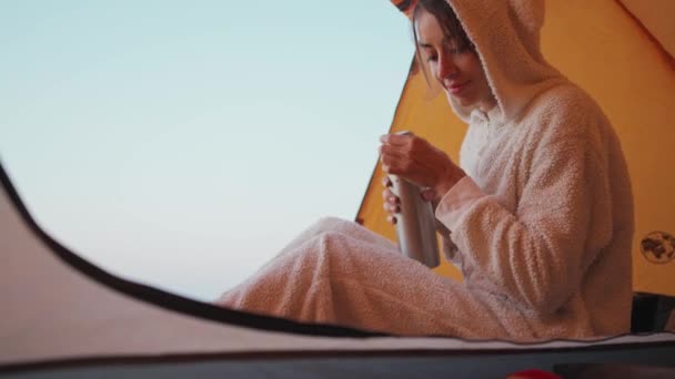 Ładna młoda kobieta w zabawnej piżamie króliczka siedzi w namiocie i nalewa ciepłą kawę z termosu do kubka. — Wideo stockowe
