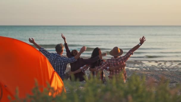 Вид сзади четыре лучших друга сидят на закате на пляже перед морем и поднимают руки — стоковое видео