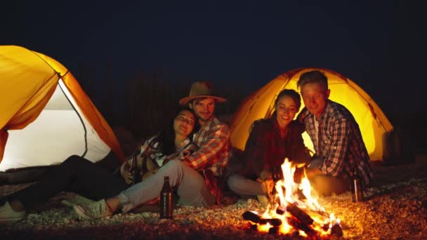 Amigos relaxando, aquecendo e se divertindo pela fogueira no acampamento com tendas na praia — Vídeo de Stock